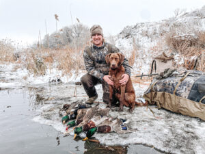 Hunter with Chesapeake Bay Retriever and ducks at edge of water in winter.